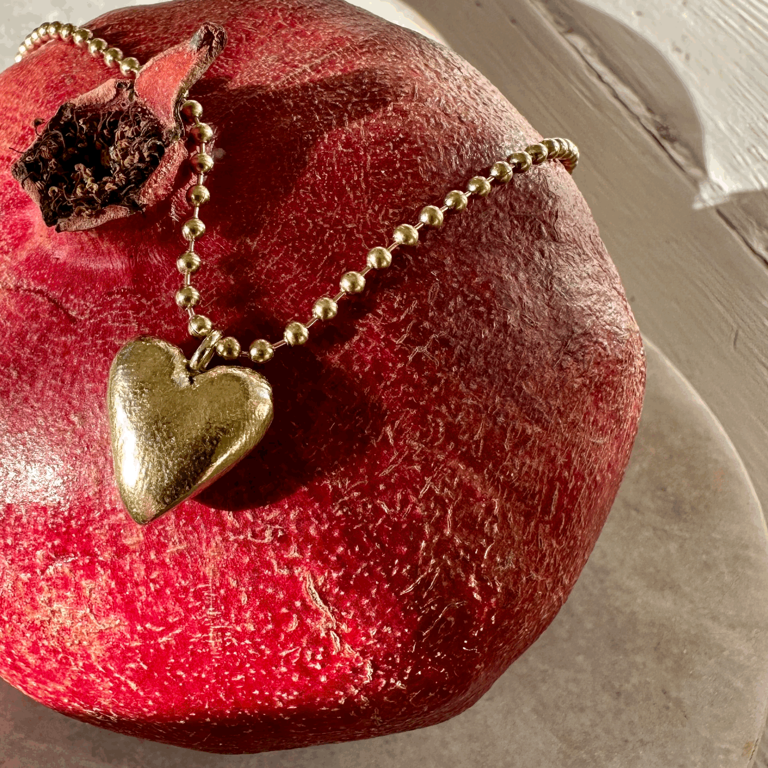 Heart Pendant: A hand carved solid recycled brass heart charm on a long brass ball chain, pictured on a pomegranate.