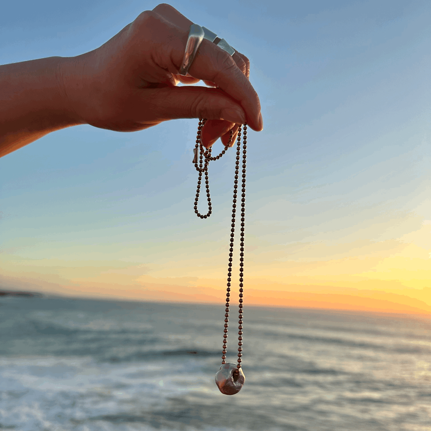 A large solid silver Hag Stone pendant held up against the backdrop of the sea.